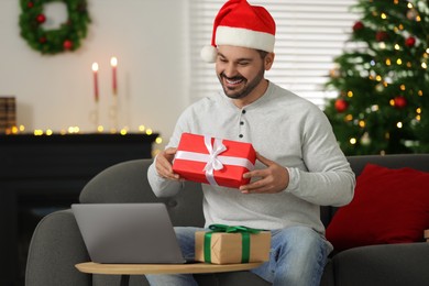 Photo of Celebrating Christmas online with exchanged by mail presents. Happy man in Santa hat with gift box during video call on laptop at home