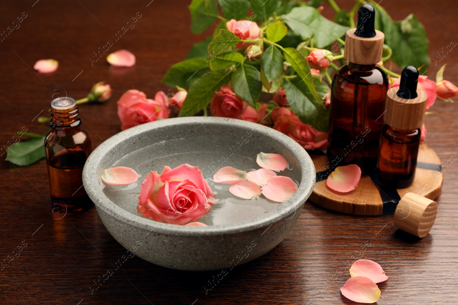 Photo of Bowl of water, bottles with essential oil and beautiful rose petals on wooden table. Aromatherapy treatment