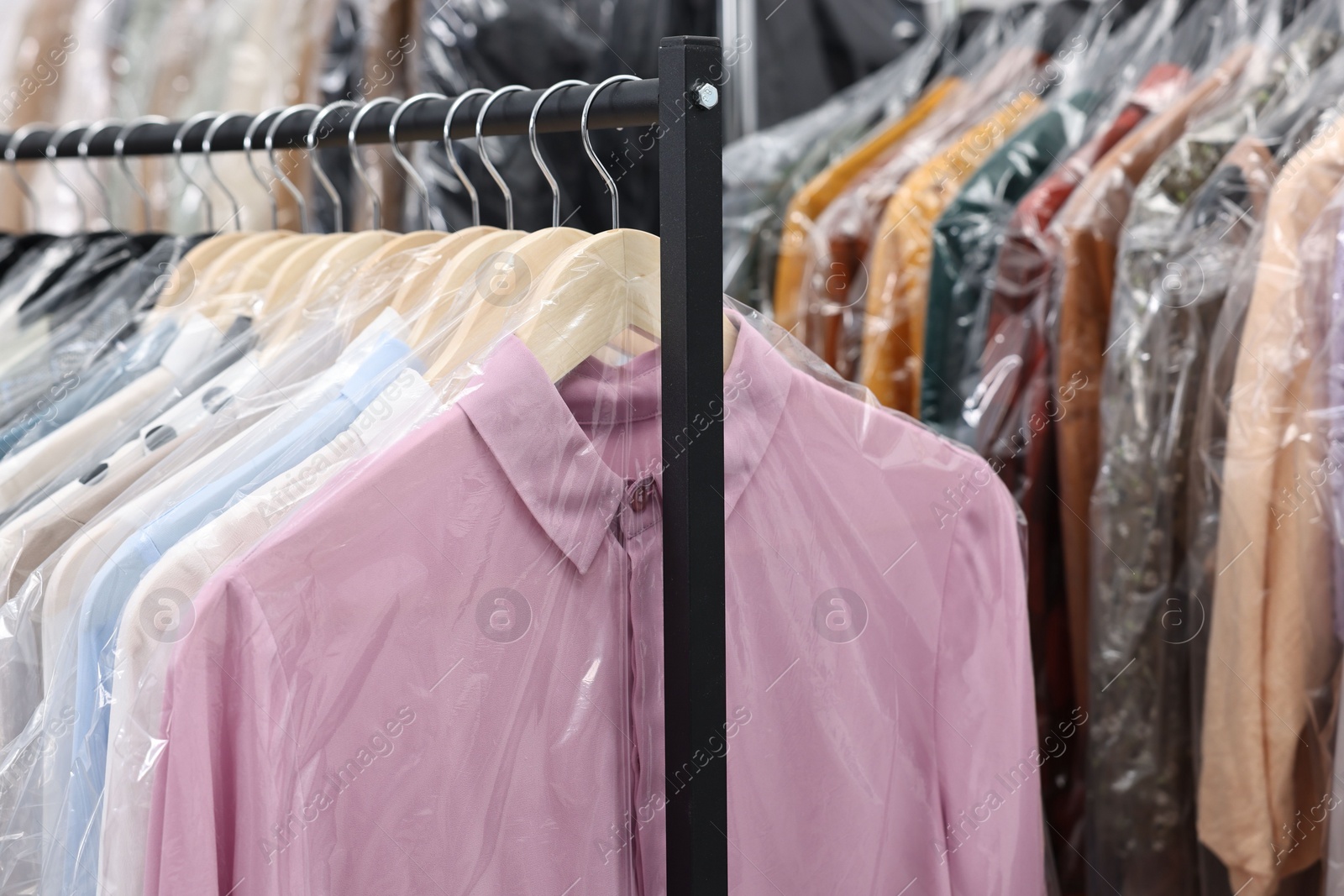 Photo of Dry-cleaning service. Many different clothes in plastic bags hanging on rack indoors, closeup