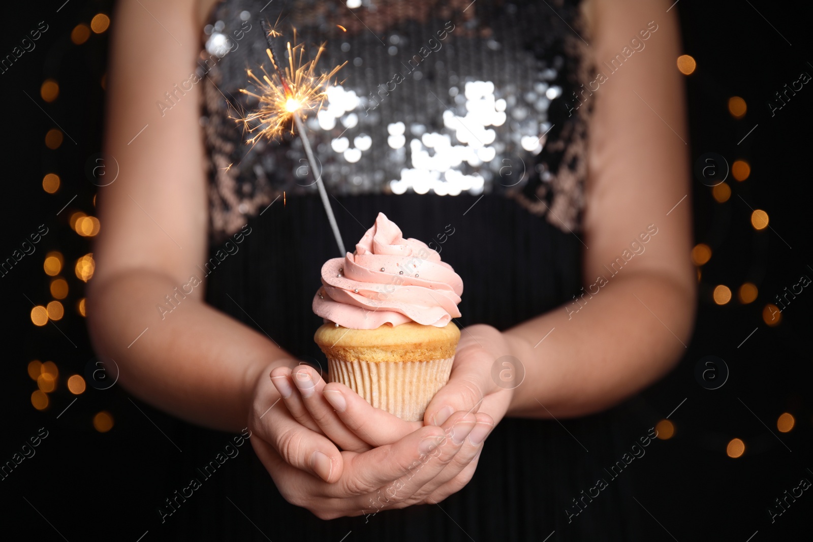 Photo of Woman holding birthday cupcake with sparkler on blurred background, closeup