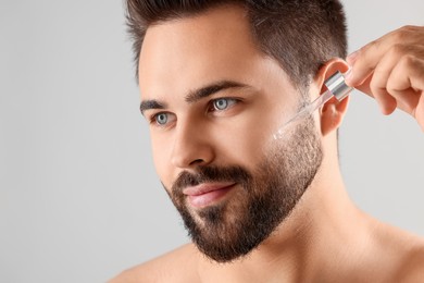 Photo of Handsome man applying cosmetic serum onto face on light grey background, closeup