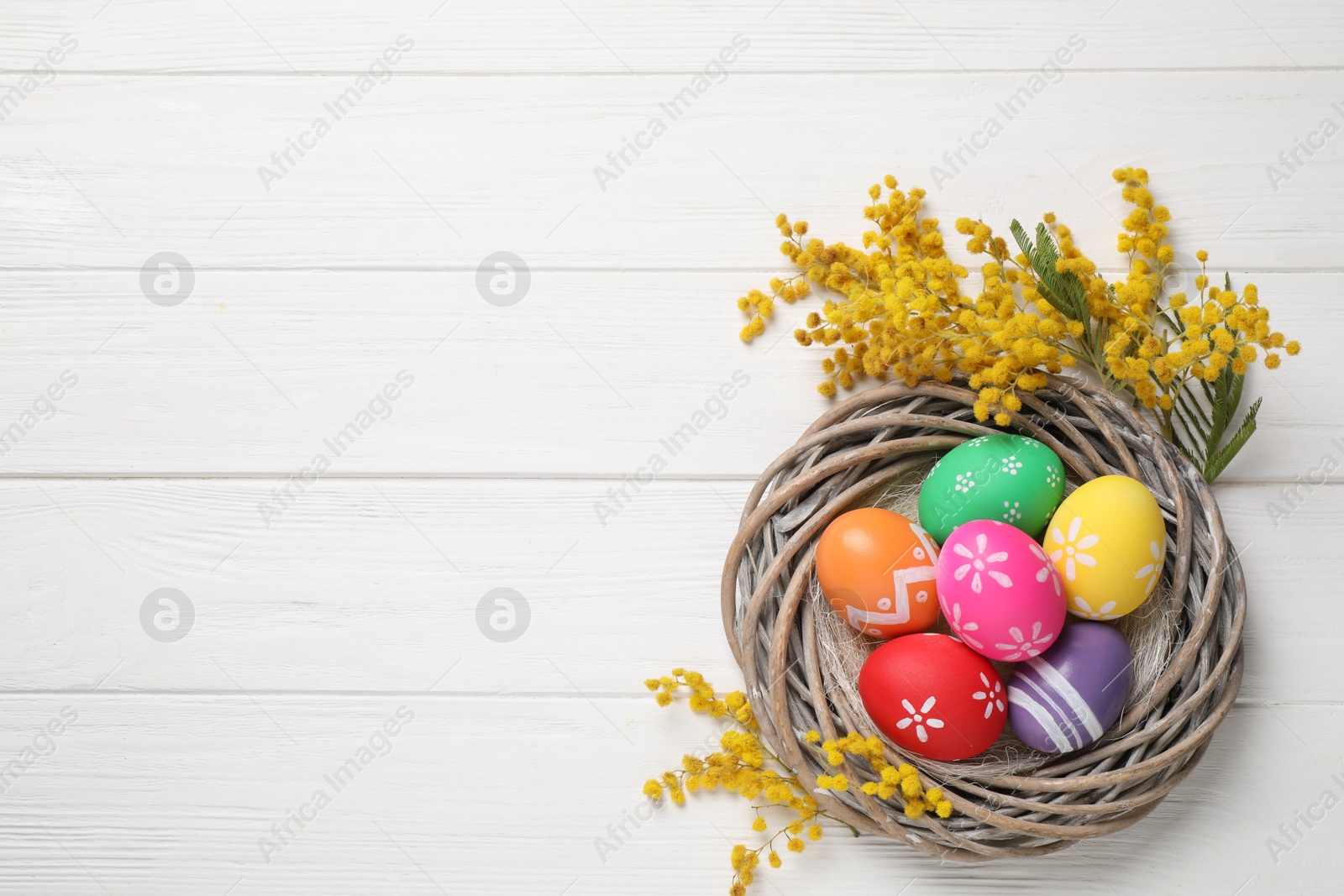 Photo of Colorful Easter eggs in decorative nest and mimosa flowers on white wooden background, flat lay. Space for text