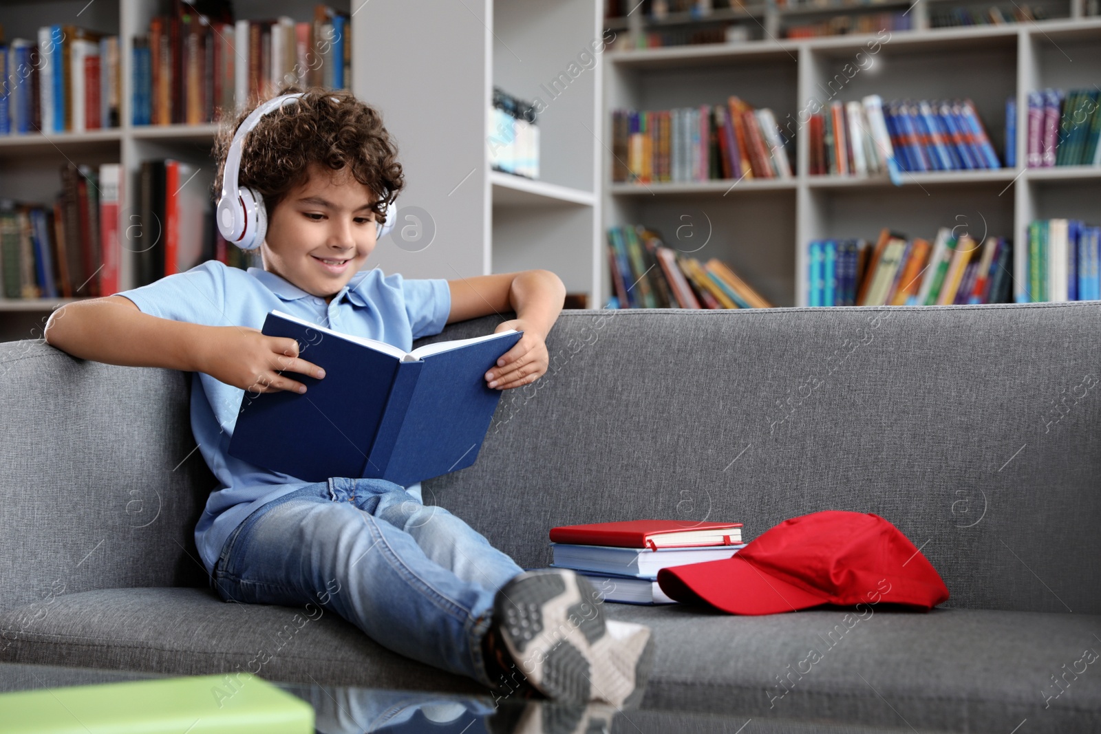 Photo of Cute little boy in headphones reading book on sofa in library