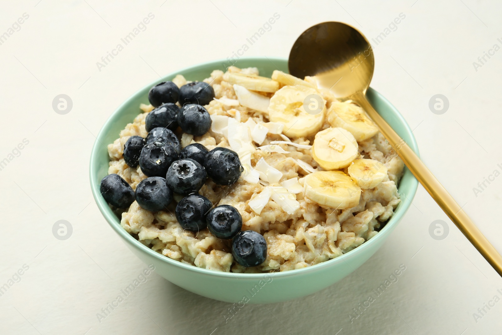 Photo of Tasty oatmeal with banana, blueberries, coconut flakes and honey served in bowl on beige table