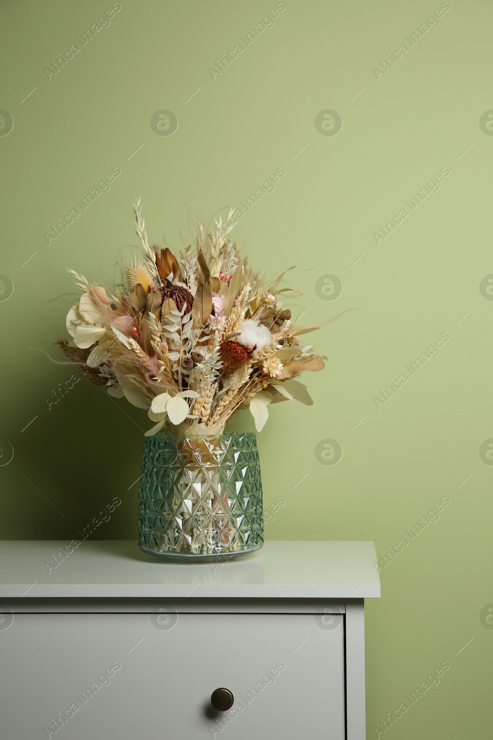 Photo of Beautiful dried flower bouquet in glass vase on white chest of drawers near olive wall