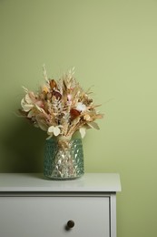 Beautiful dried flower bouquet in glass vase on white chest of drawers near olive wall