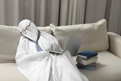 Photo of Overworked ghost. Man in white sheet with laptop and books on sofa at home