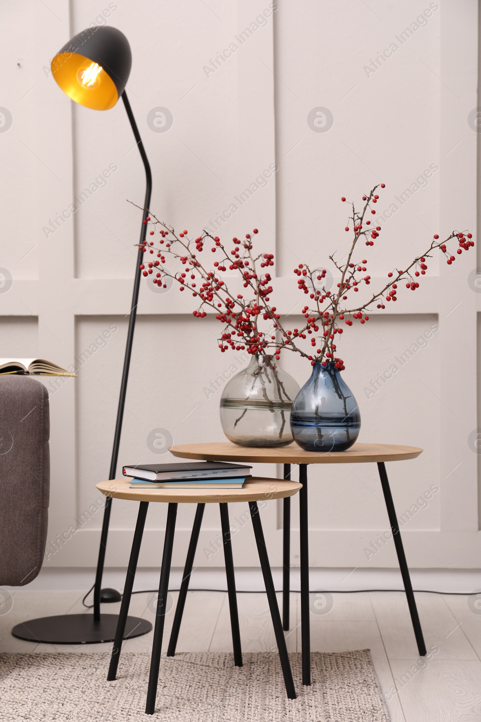 Photo of Hawthorn branches with red berries in vases and lamp near light wall indoors. Interior design
