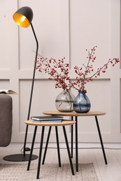 Hawthorn branches with red berries in vases and lamp near light wall indoors. Interior design