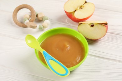 Photo of Baby food. Puree of apples in bowl, spoon and toy on white wooden table