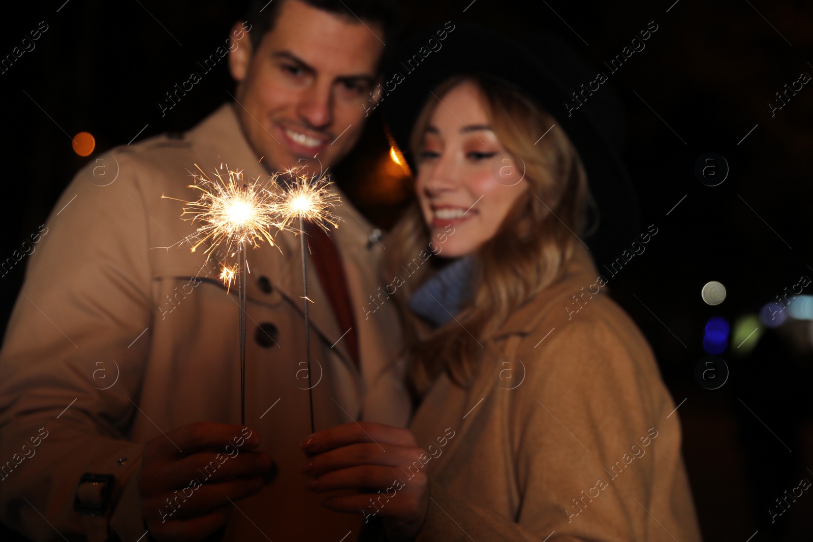 Photo of Couple in warm clothes holding burning sparklers at night, focus on fireworks