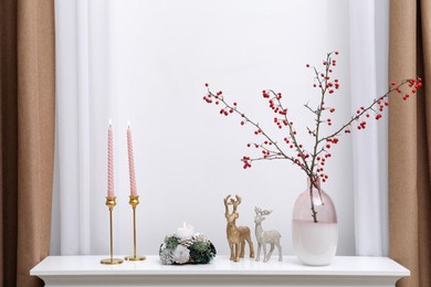 Photo of Hawthorn branches with red berries in vase, candles and deer figures on white table near window indoors