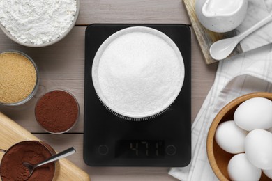 Electronic scales with sugar and different products on light wooden table, flat lay