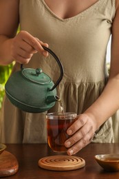 Woman pouring freshly brewed tea from teapot into cup at wooden table, closeup. Traditional ceremony