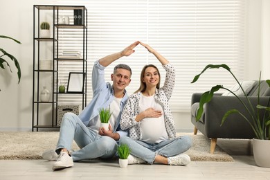 Photo of Young family housing concept. Pregnant woman with her husband forming roof with their hands while sitting on floor at home
