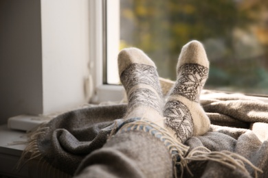 Woman in knitted socks relaxing on plaid near window at home, closeup