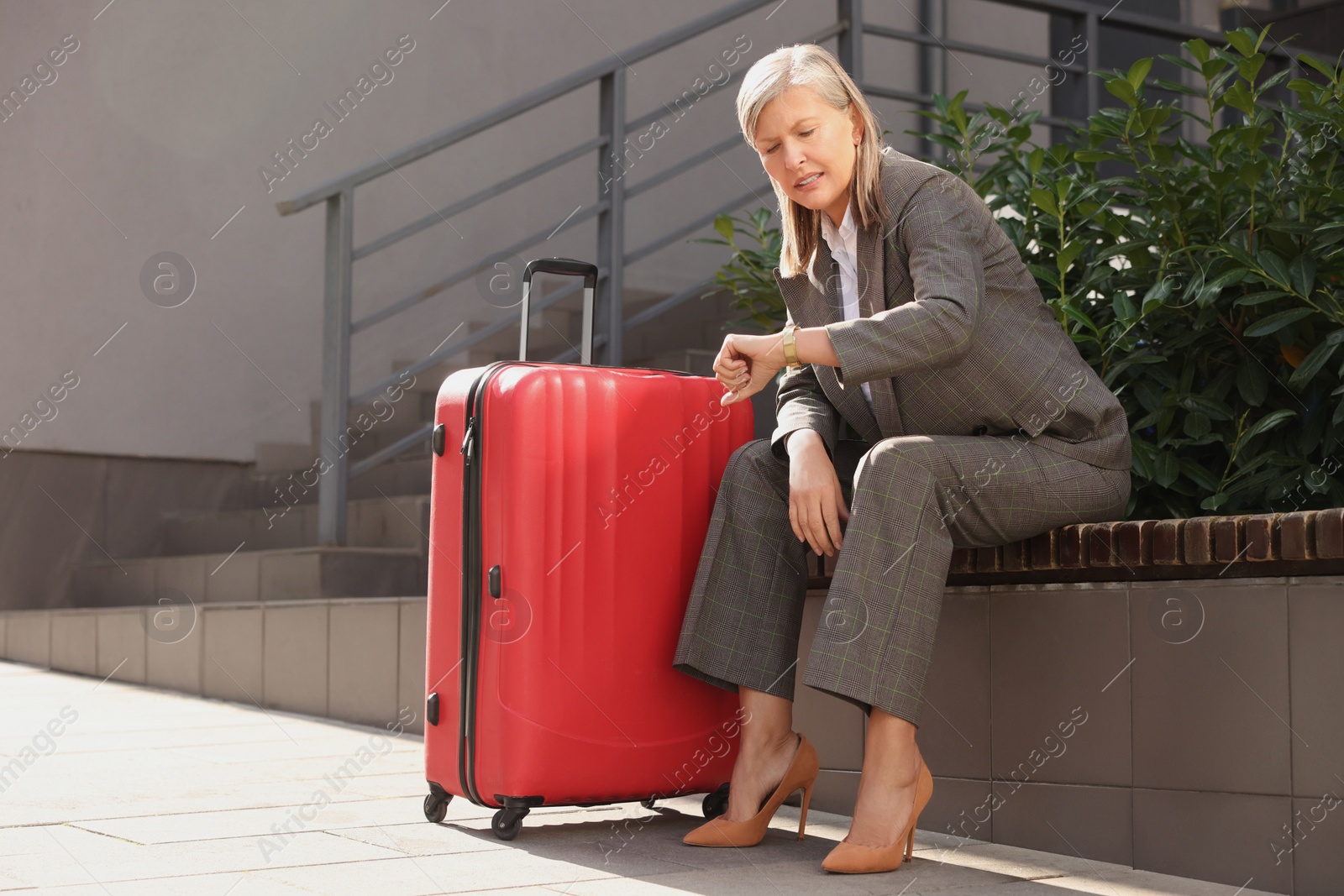 Photo of Being late. Worried senior businesswoman with red suitcase looking at her watch while sitting on bench outdoors