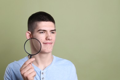 Young man with acne problem holding magnifying glass on olive background. Space for text