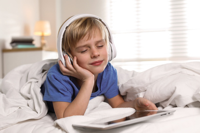 Photo of Cute little boy with headphones and tablet listening to audiobook in bed at home