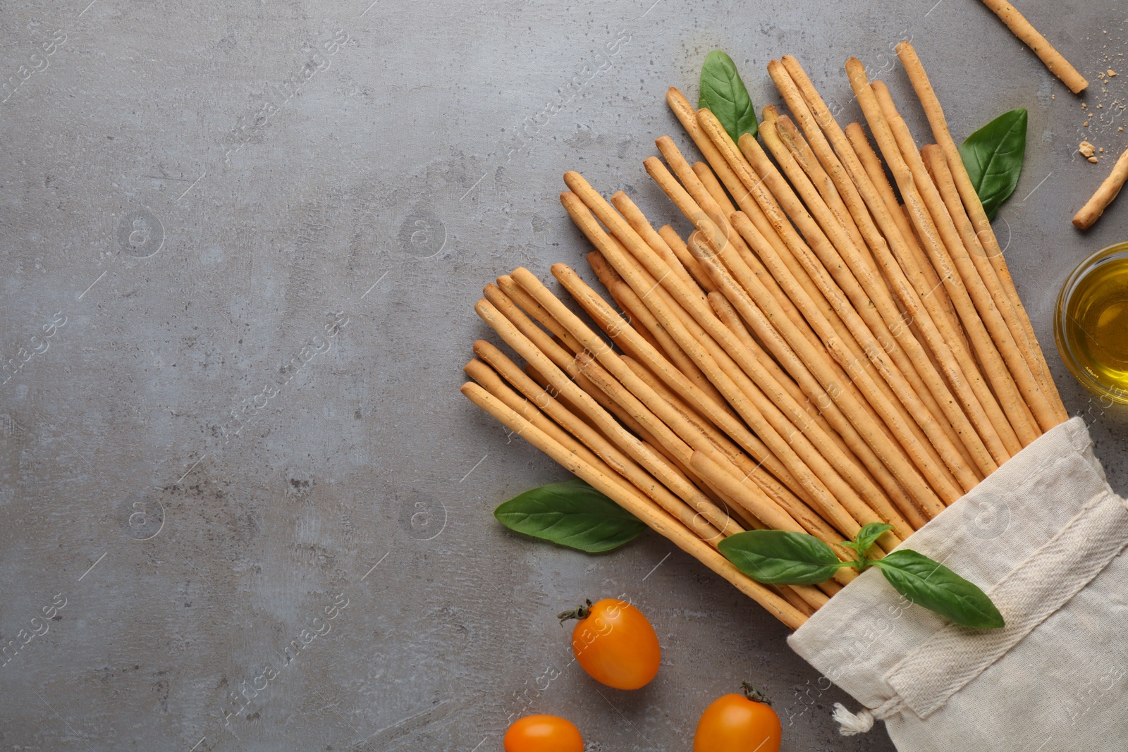 Photo of Delicious grissini sticks, basil leaves, cherry tomatoes and oil on grey table, flat lay. Space for text