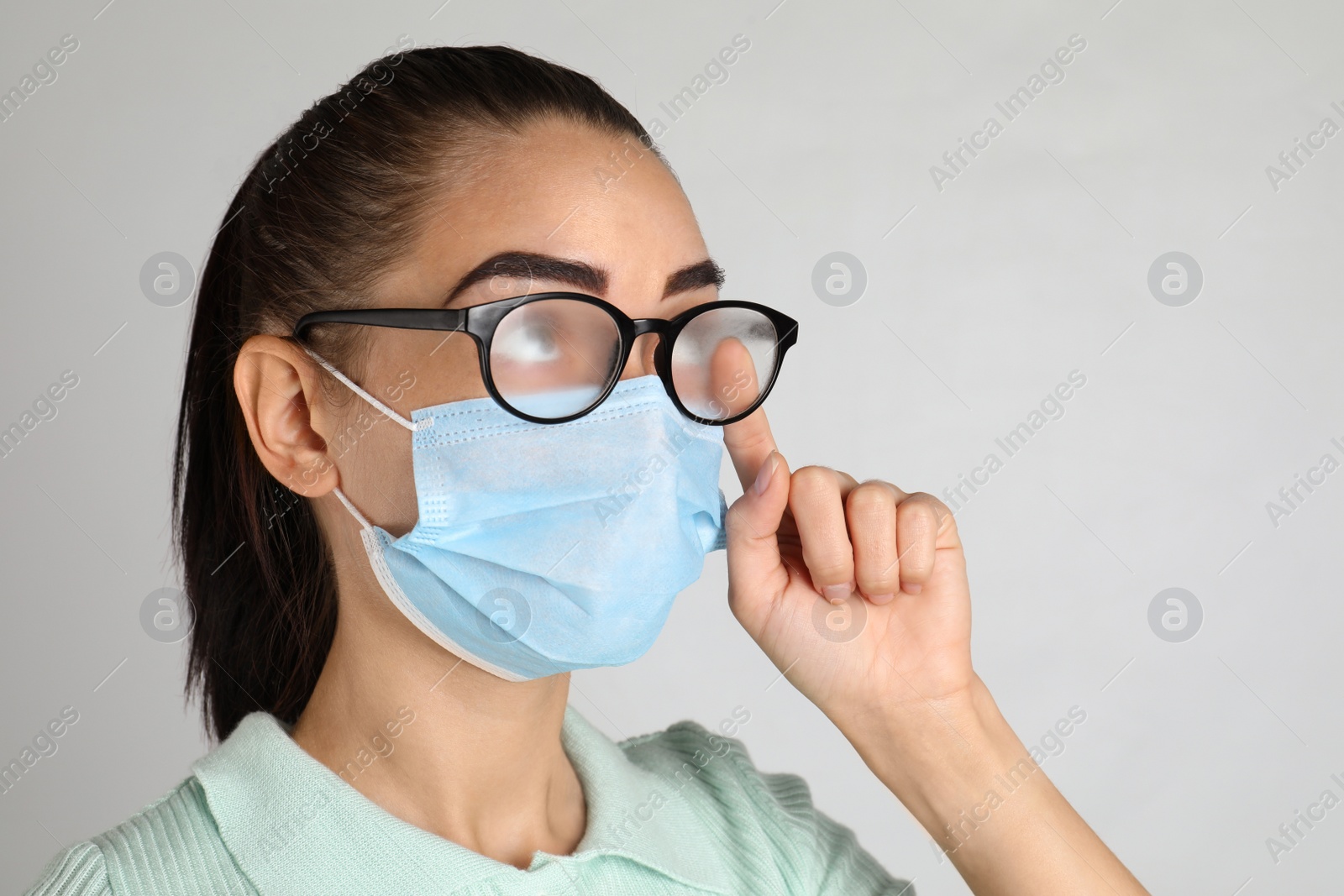 Photo of Woman wiping foggy glasses caused by wearing medical mask on light background