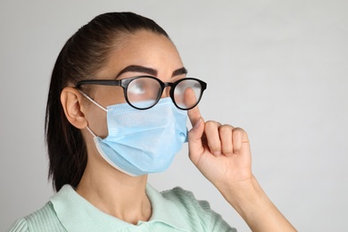 Photo of Woman wiping foggy glasses caused by wearing medical mask on light background