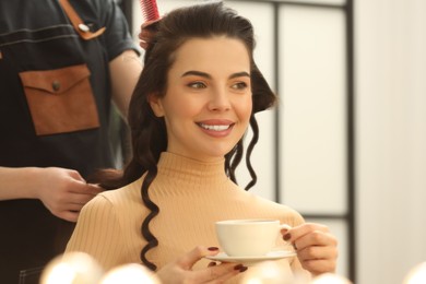 Photo of Hair styling. Professional hairdresser working with smiling woman while she drinking coffee in salon, closeup. Space for text