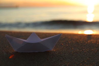 Photo of White paper boat on sand near sea at sunset, closeup. Space for text