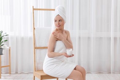 Beautiful young woman applying body cream onto shoulder indoors