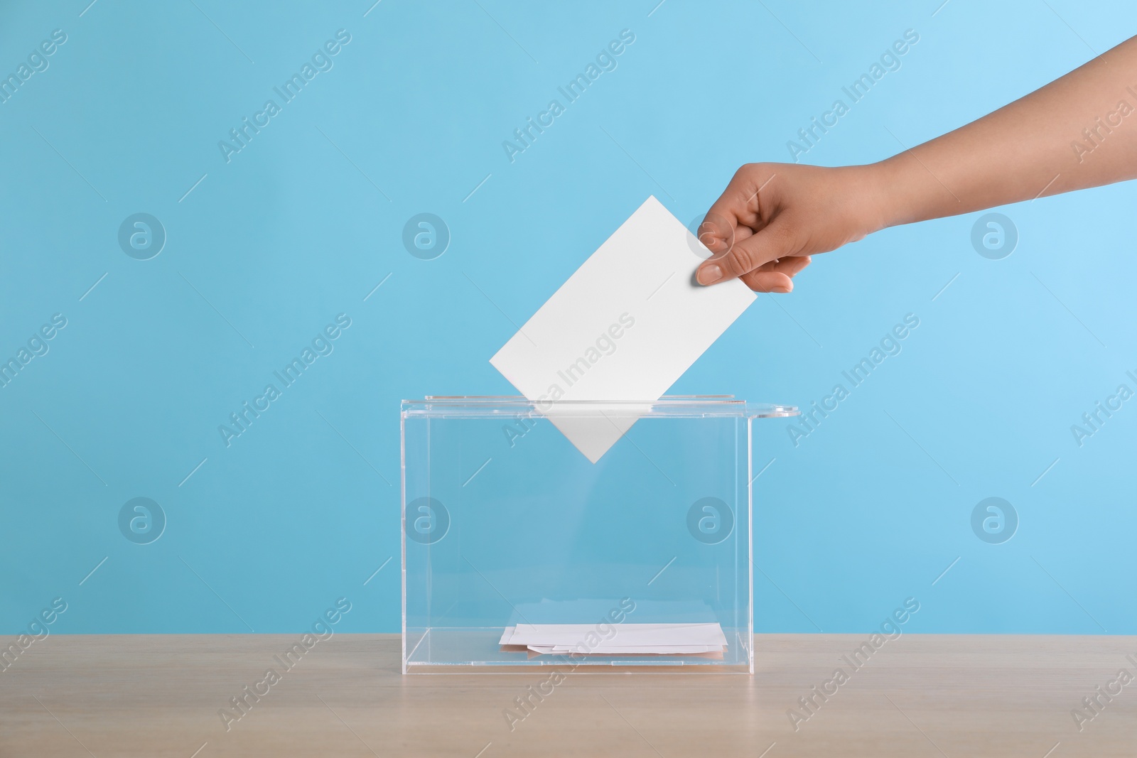 Photo of Woman putting her vote into ballot box on wooden table against light blue background, closeup