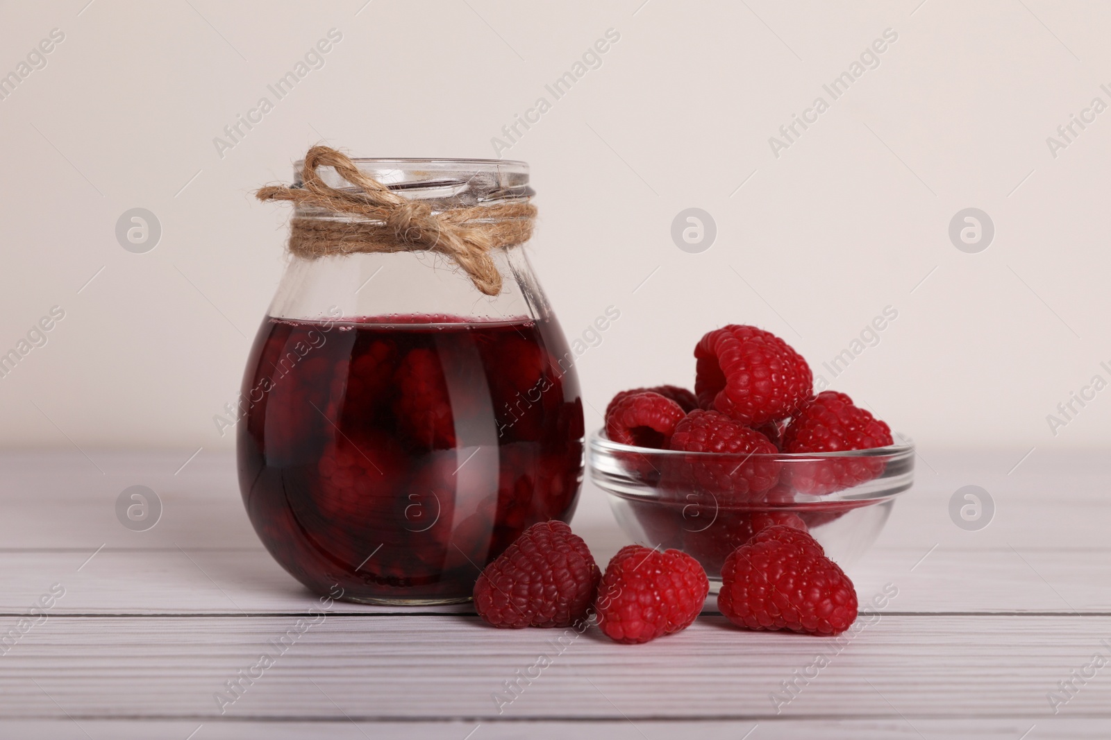 Photo of Jar of tasty canned raspberry jam and fresh berries in glass bowl on white wooden table