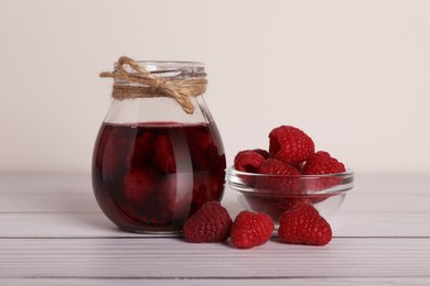 Photo of Jar of tasty canned raspberry jam and fresh berries in glass bowl on white wooden table