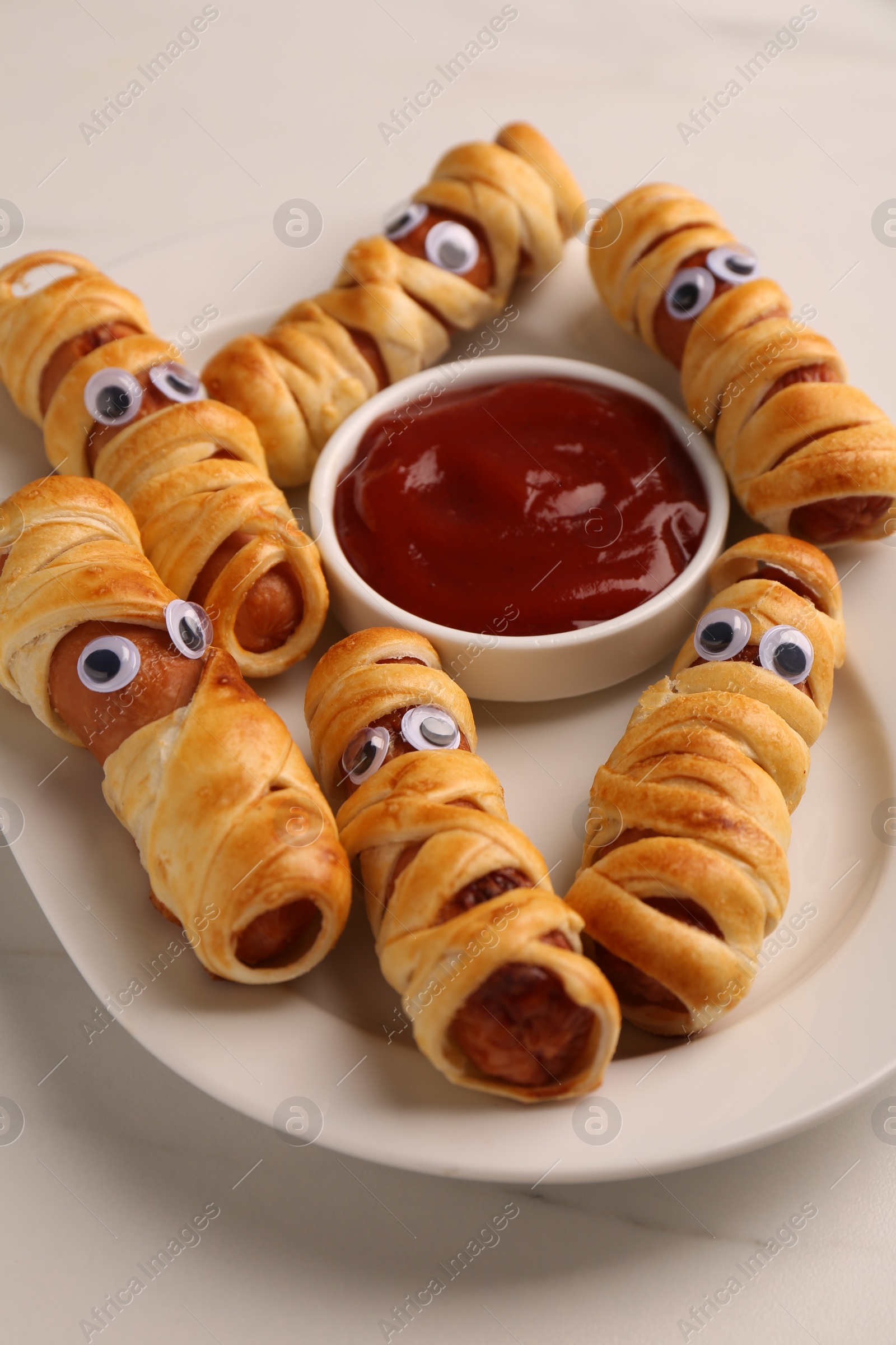 Photo of Plate with tasty sausage mummies for Halloween party and ketchup on white table, closeup