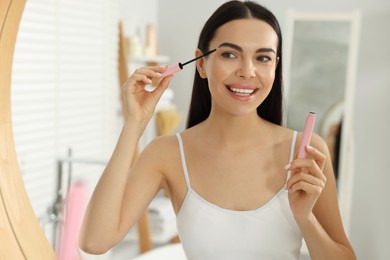 Beautiful young woman applying mascara near mirror in bathroom