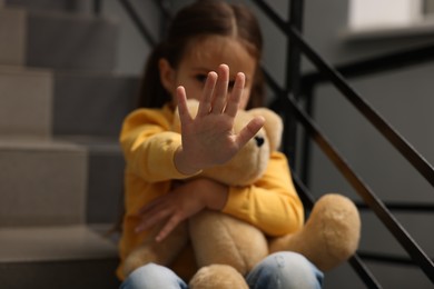 Photo of Child abuse. Little girl with teddy bear doing stop gesture indoors, selective focus