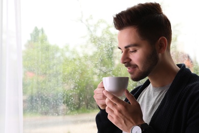 Happy handsome man with cup of coffee near window on rainy day