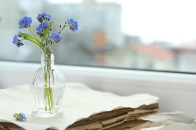 Beautiful blue forget-me-not flowers in glass bottle on stack of old paper near window. Space for text