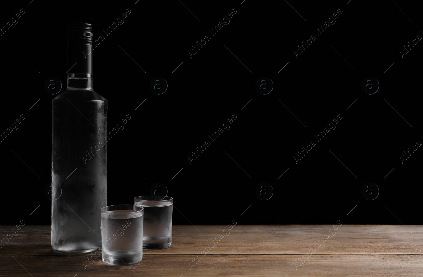 Photo of Bottle of vodka and shot glasses on wooden table against black background. Space for text