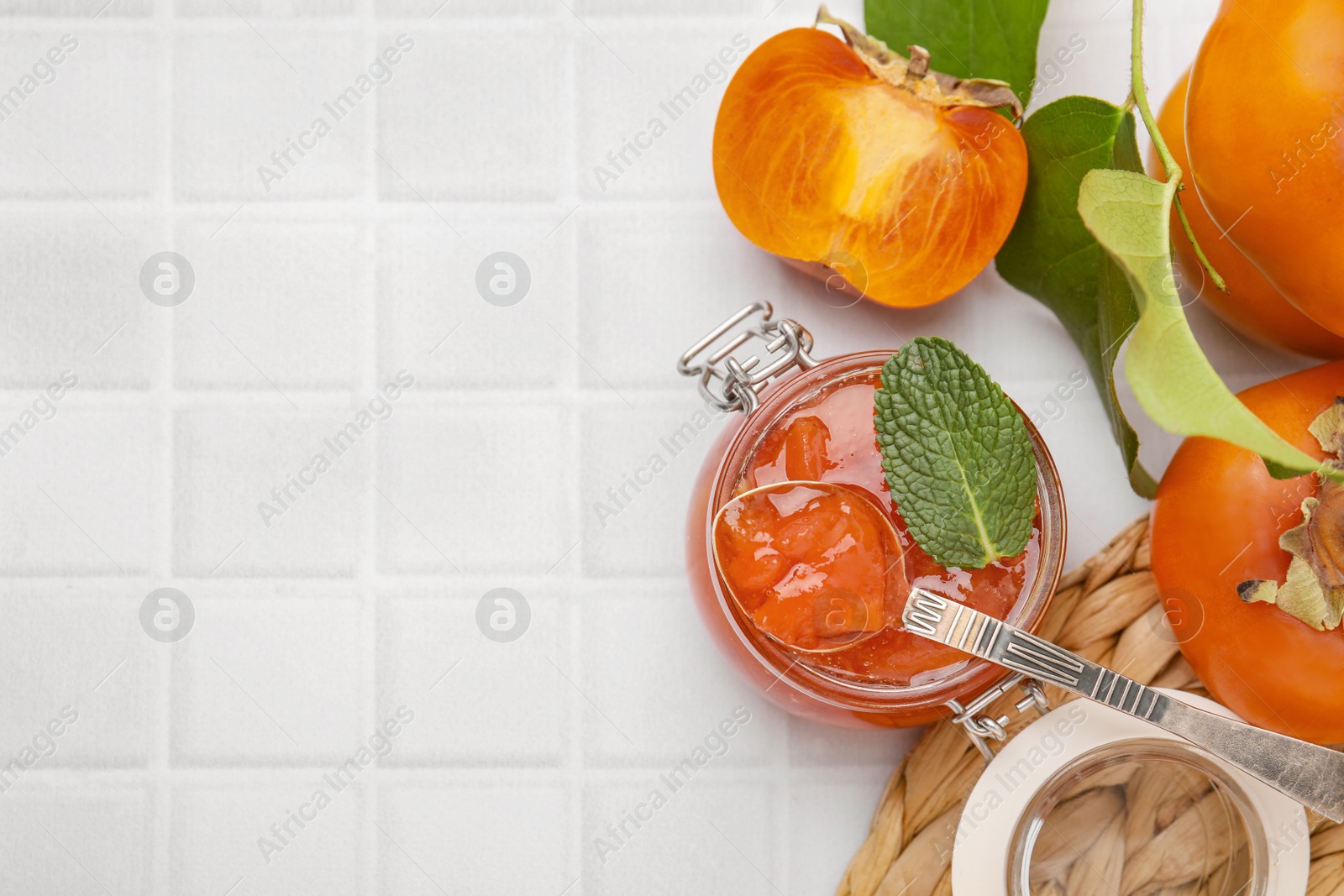 Photo of Jar and spoon of tasty persimmon jam, ingredients on white tiled table, flat lay. Space for text