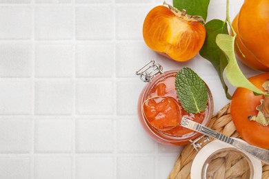 Photo of Jar and spoon of tasty persimmon jam, ingredients on white tiled table, flat lay. Space for text