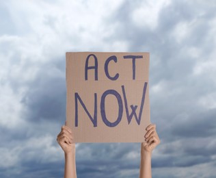 Image of Protestor holding placard with text Act Now against cloudy sky, closeup. Climate strike