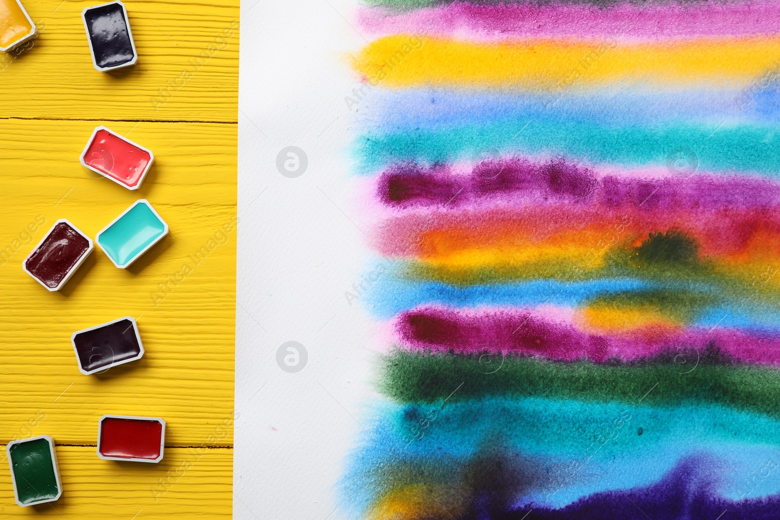 Photo of Bright abstract watercolor painting and colorful paints on yellow wooden table, flat lay