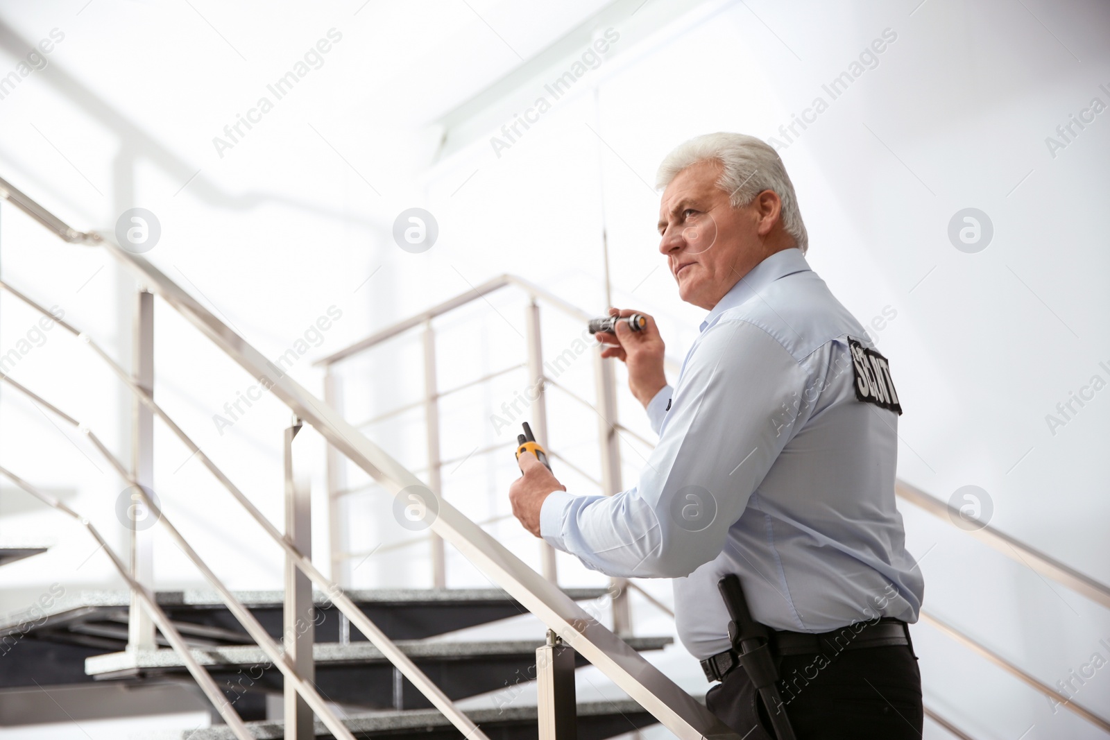 Photo of Professional security guard with portable radio set and flashlight on stairs indoors