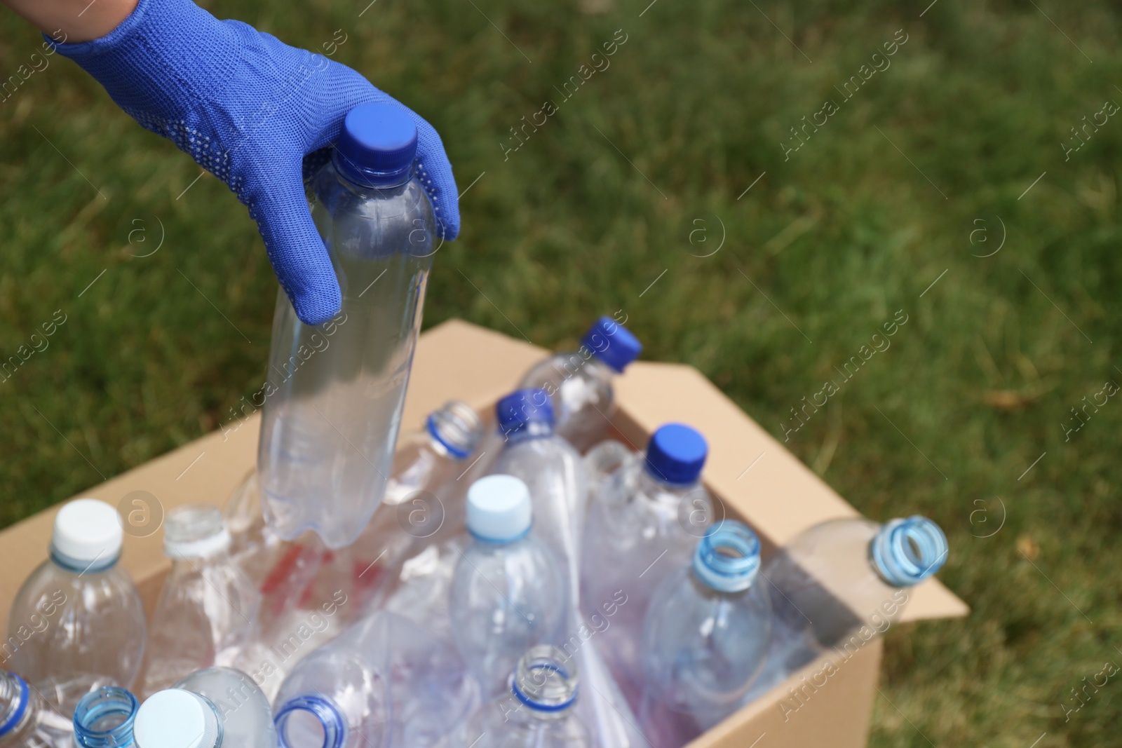 Photo of Woman in gloves putting used plastic bottle into cardboard box outdoors, closeup. Recycling problem