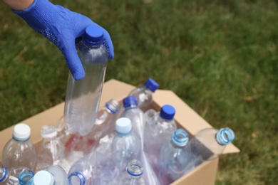 Woman in gloves putting used plastic bottle into cardboard box outdoors, closeup. Recycling problem