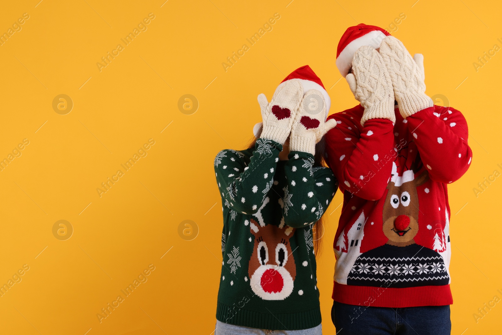 Photo of Couple in Christmas sweaters and Santa hats covering faces with hands in knitted mittens on orange background. Space for text