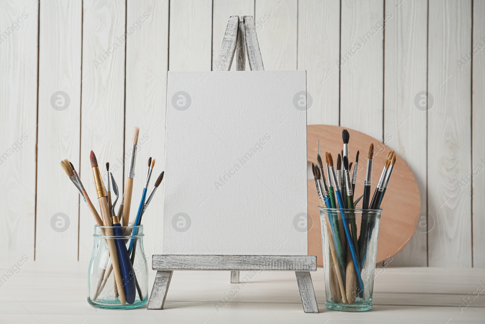 Photo of Easel with blank canvas and brushes on white wooden table