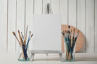 Photo of Easel with blank canvas and brushes on white wooden table