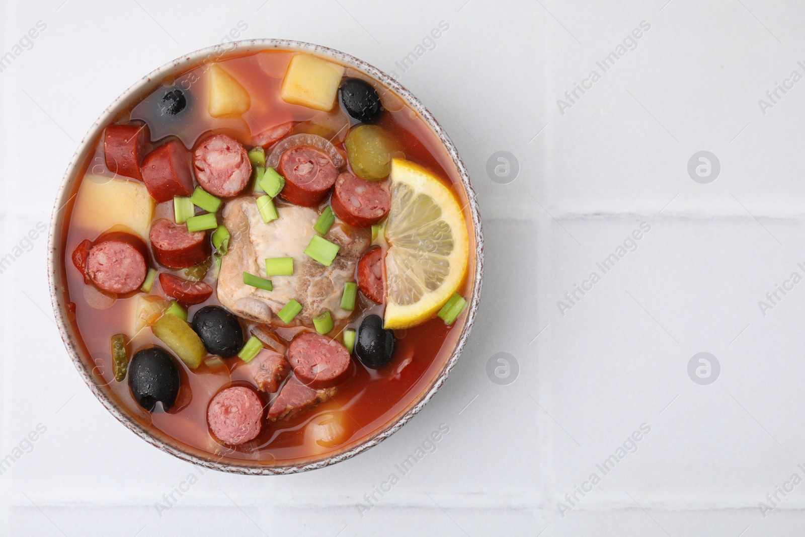 Photo of Meat solyanka soup with thin dry smoked sausages in bowl on white tiled table, top view. Space for text