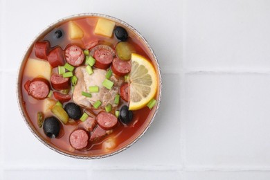 Photo of Meat solyanka soup with thin dry smoked sausages in bowl on white tiled table, top view. Space for text